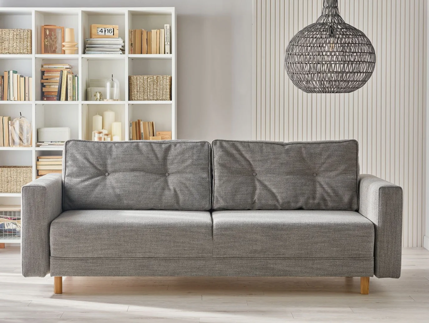 Szara sofa na tle białego regału z książkami i lamelowej ściany
